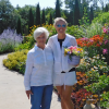 Belinda Jensen and her mom, Jackie, standing next to lush garden in full bloom on a sunny spring day; Belinda smiles as she holds a patio planter
