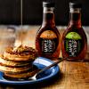 A stack of homemade waffles are drizzled with maple syrup; bottles of Kowalski's Signature 100% Pure and Organic Maple Syrup sit on the table in the background